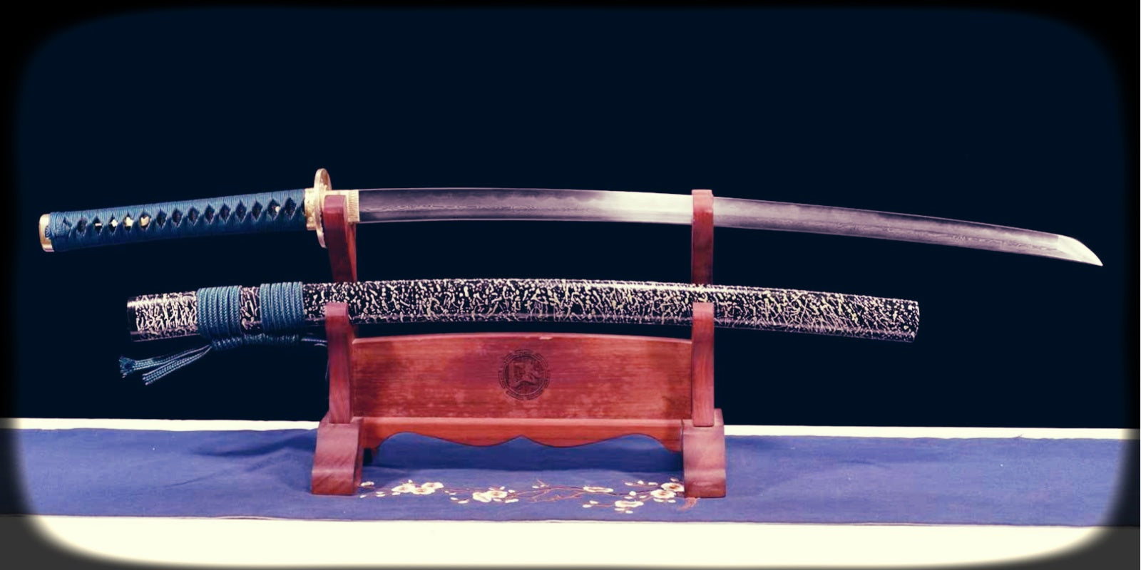 Clay-tempered samurai sword displayed on a wooden stand with visible hamon, blue tsuka wrap, and a textured dark saya, illustrating the kind of blade surface and temper line discussed in the guide to spotting real clay tempering.