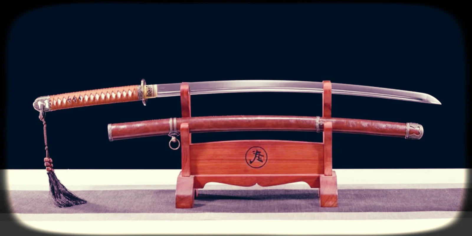 A katana-style sword with a polished curved blade and brown handle displayed above its matching brown saya on a wooden stand against a dark background.
