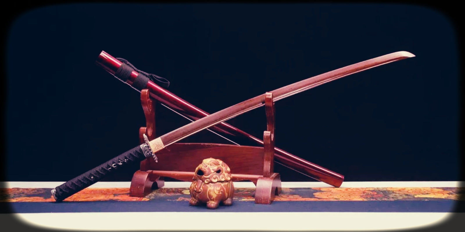Damascus folded samurai sword with a red-toned blade, black handle wrap, matching red saya, and wooden display stand, showing layered steel aesthetics.